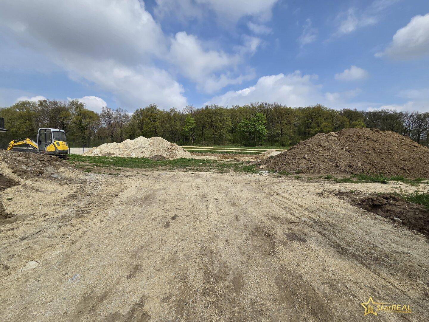 Weitläufige Baustelle mit großen Erdhügeln und einem Bagger unter blauem Himmel.