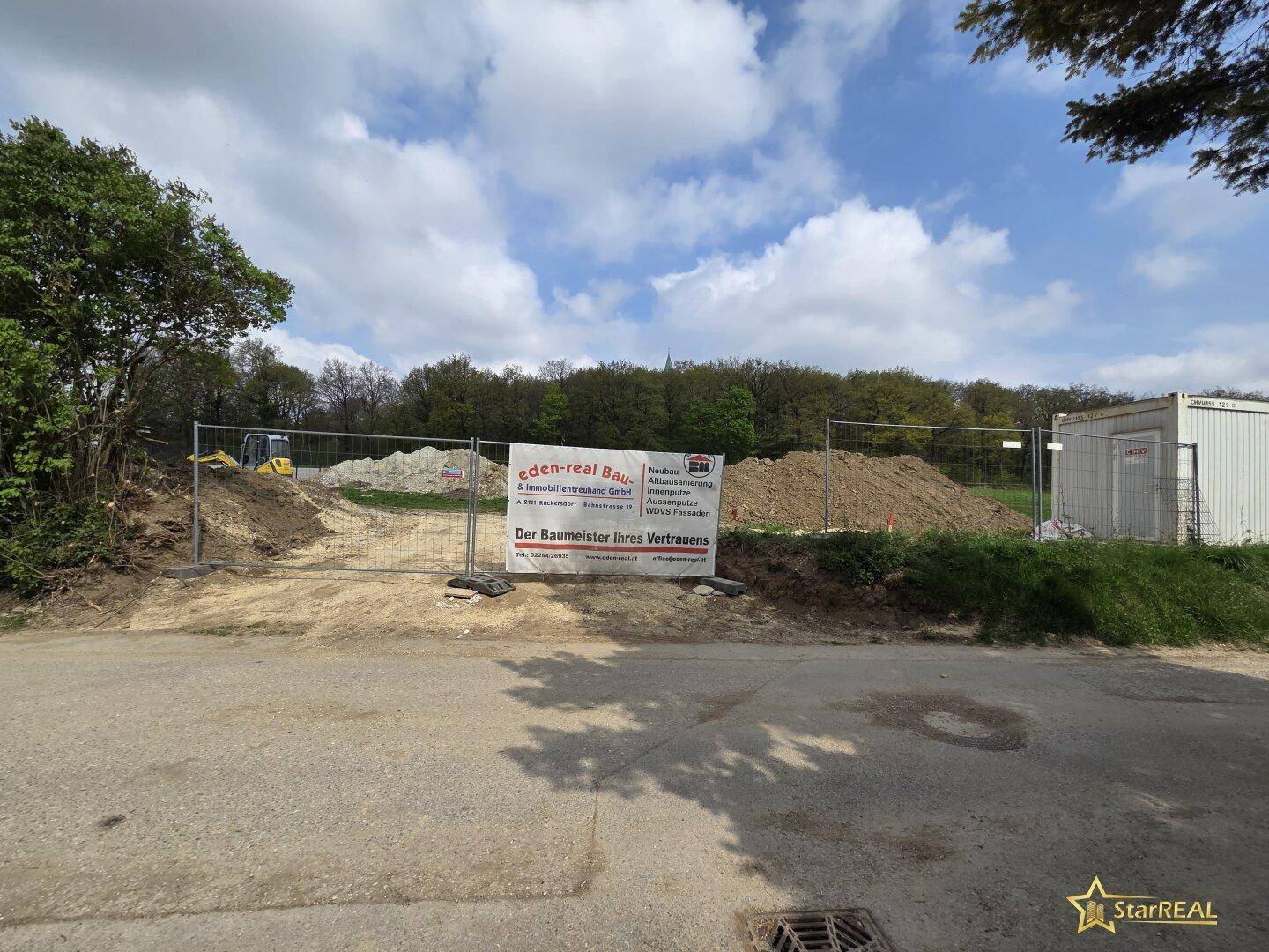 Baustelle mit Erdhügeln, einem Bagger und einem Werbebanner für Bauarbeiten.