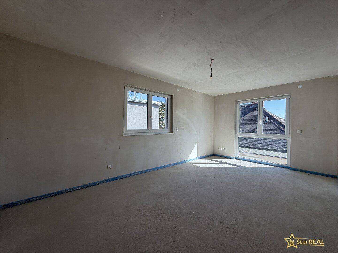 Helles Zimmer mit zwei Fenstern und Zugang zu einem Außenbereich, noch im Rohbau.