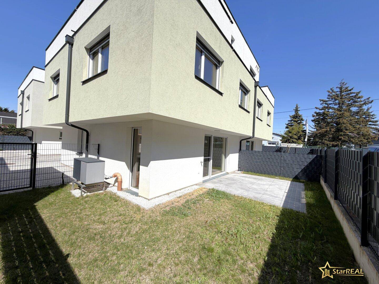 Moderne Hausfassade mit kleiner Terrasse, Rasenfläche und sichtbarer Wärmepumpe.