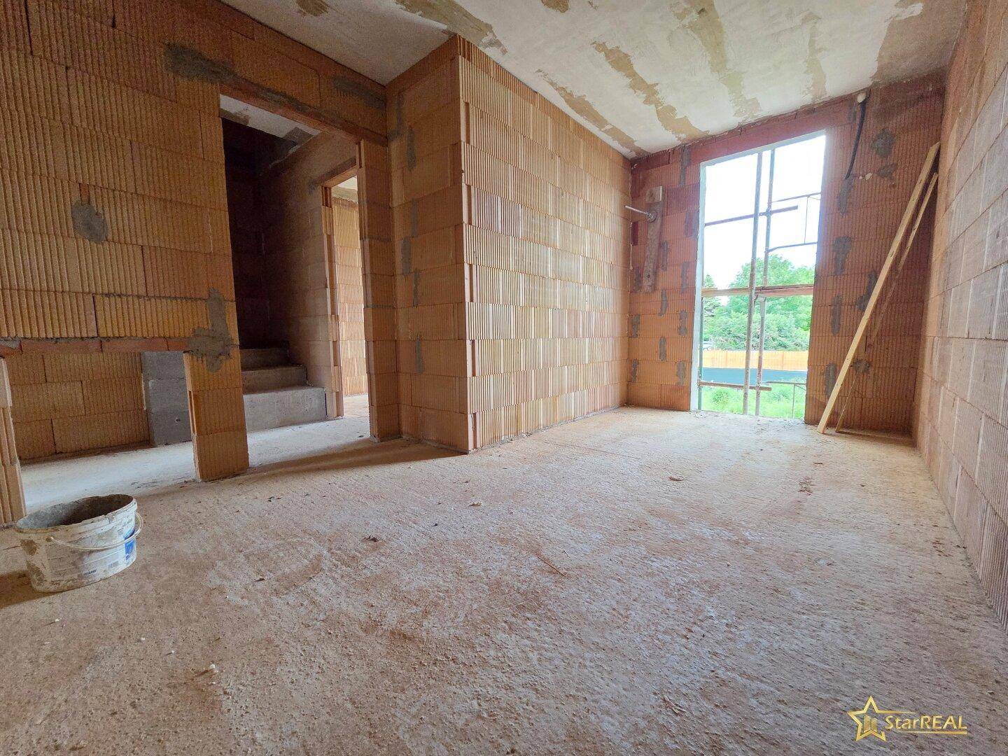 Rohbau-Raum mit hoher Fensteröffnung, Ziegelwänden und Betonboden.