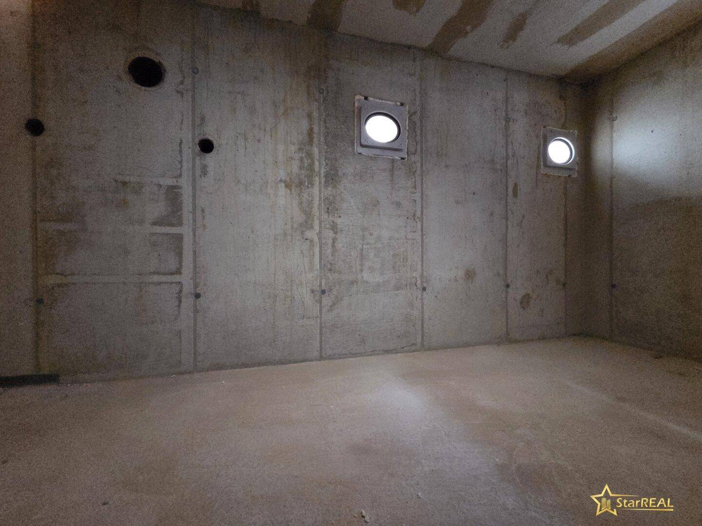 Kellerraum im Rohbau mit Betonwänden und zwei kleinen runden Fensteröffnungen.