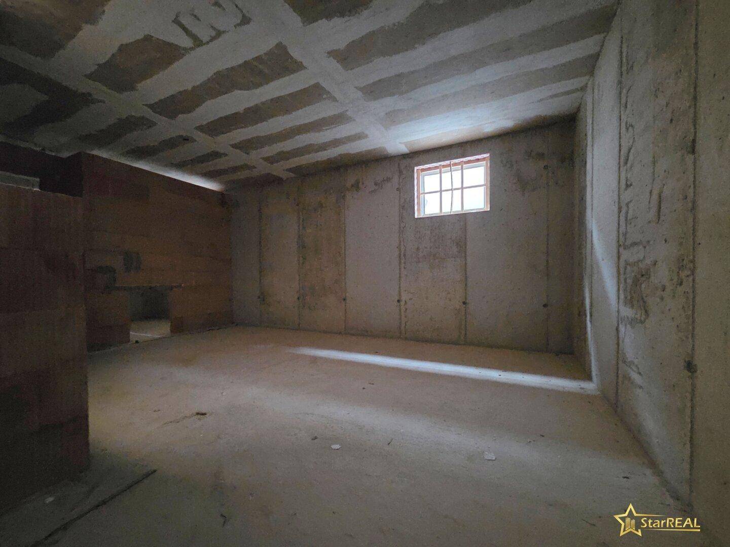 Unfertiger Kellerraum mit Betonwänden, sichtbarer Deckenstruktur und kleiner Fensteröffnung für natürliches Licht.