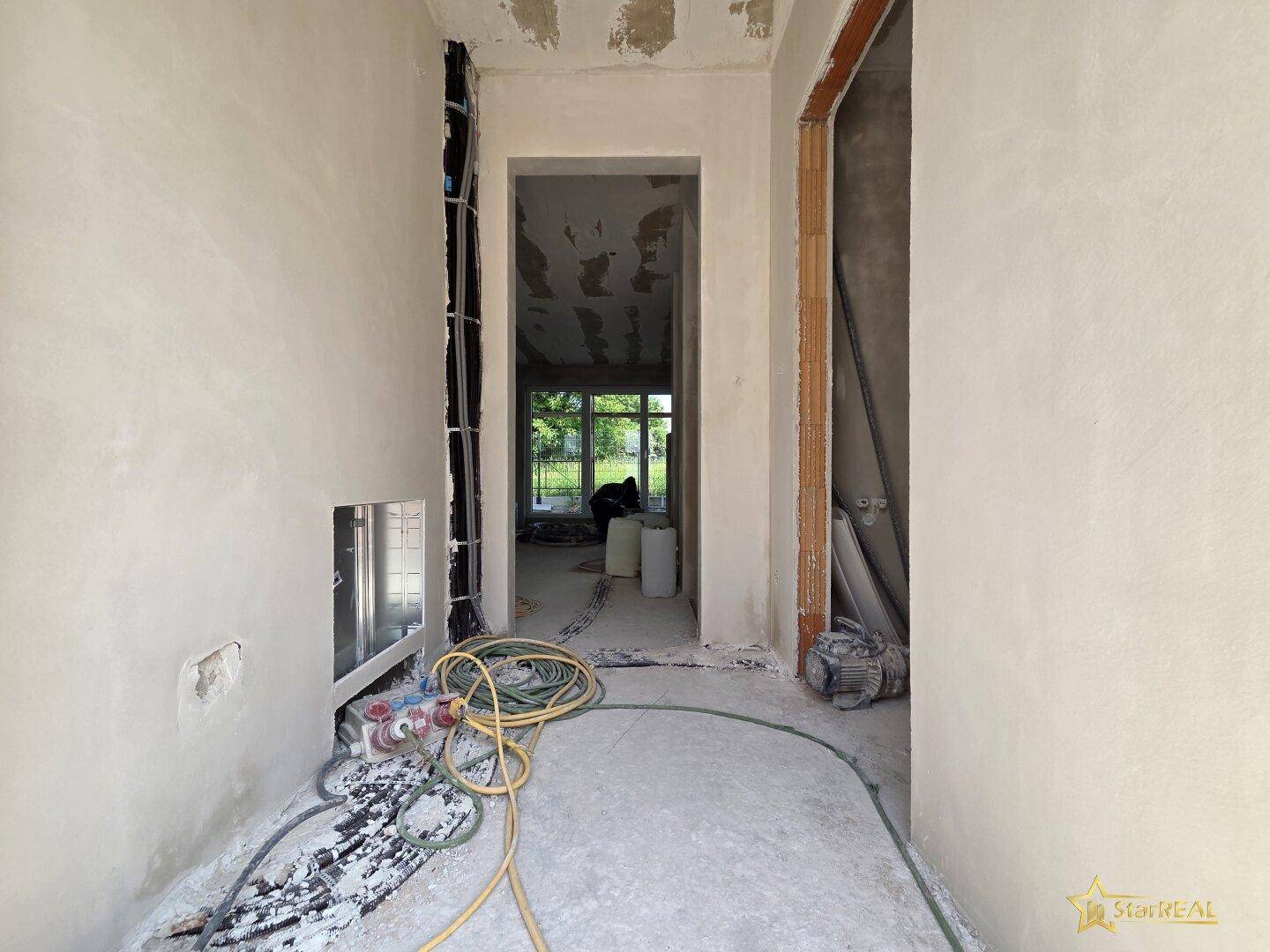 Flur im Rohbau mit sichtbaren Installationen und Durchgängen zu weiteren Räumen.