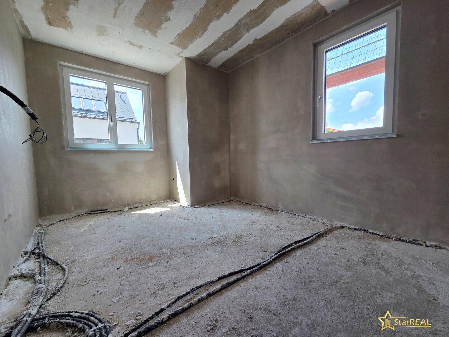 Schlafzimmer im Rohbau mit zwei Fenstern, die viel Tageslicht in den Raum lassen.