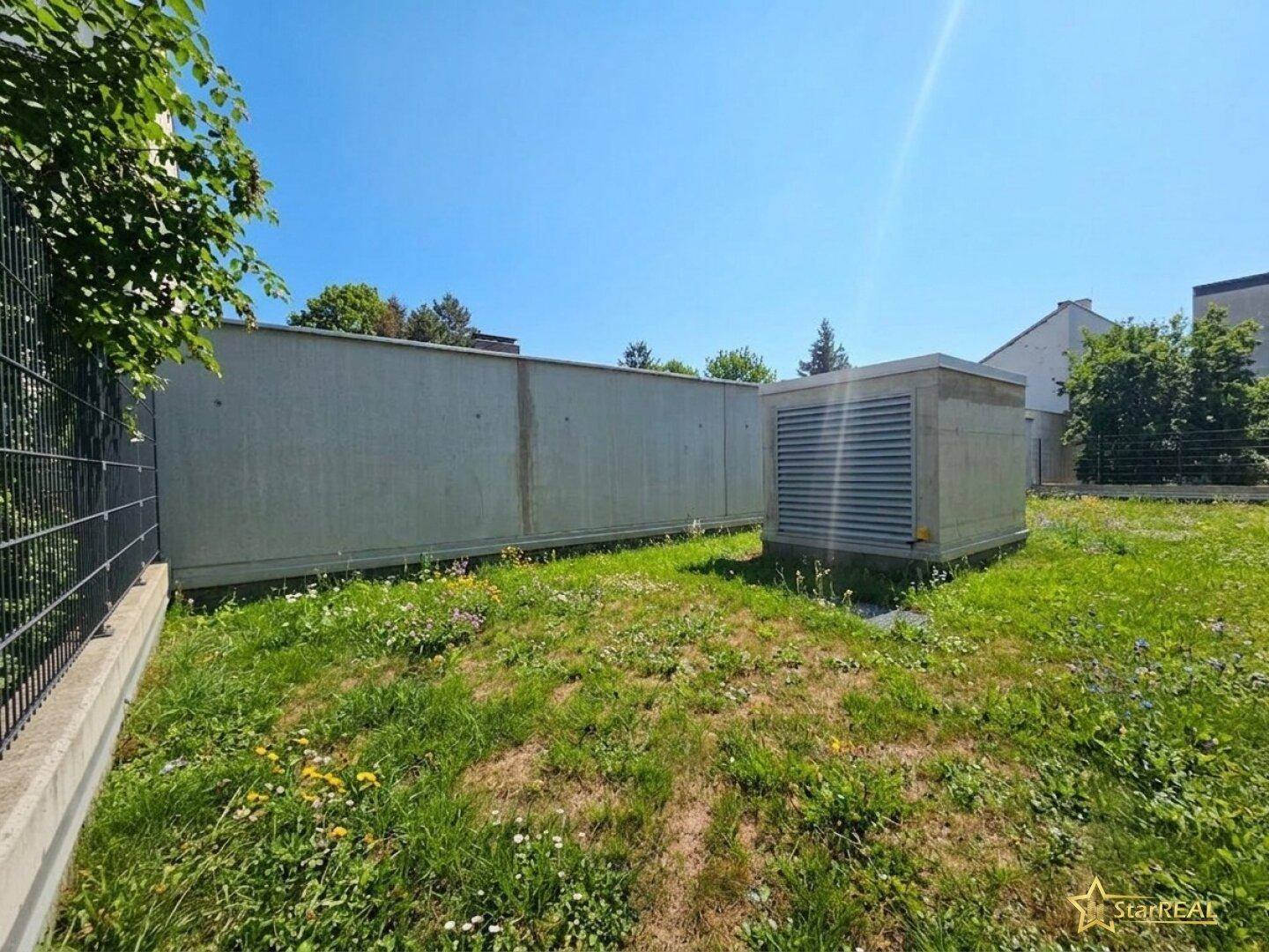 Außenbereich mit einer Grasfläche, einer Betonwand und einer technischen Anlage unter blauem Himmel.