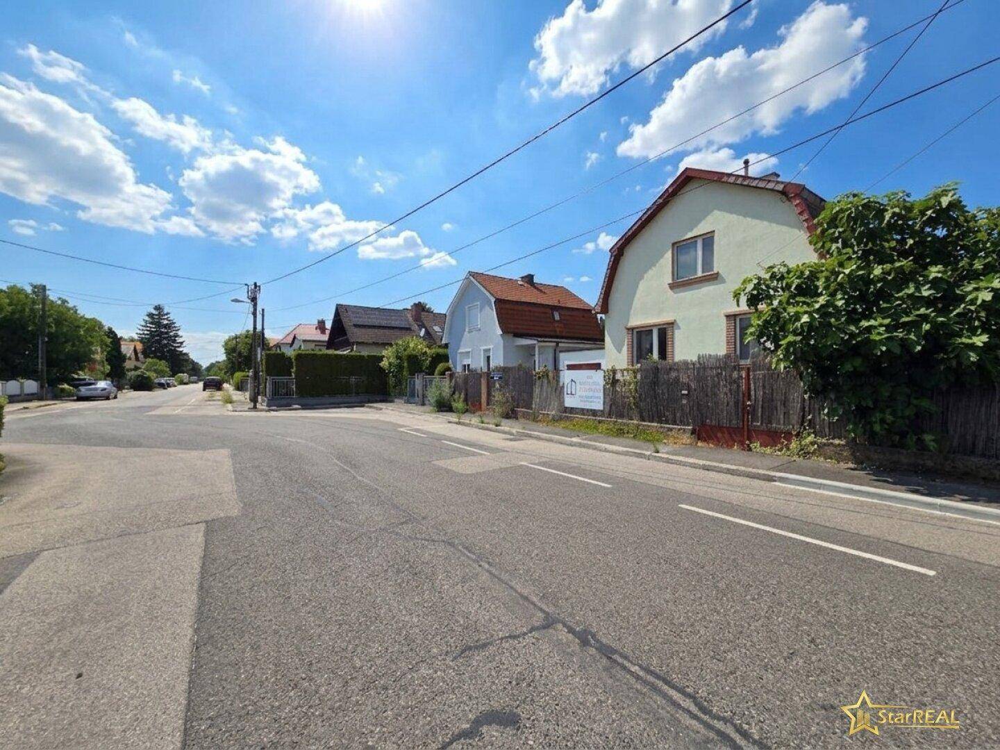 Blick entlang einer ruhigen Wohnstraße mit Einfamilienhäusern und viel Grün unter blauem Himmel.
