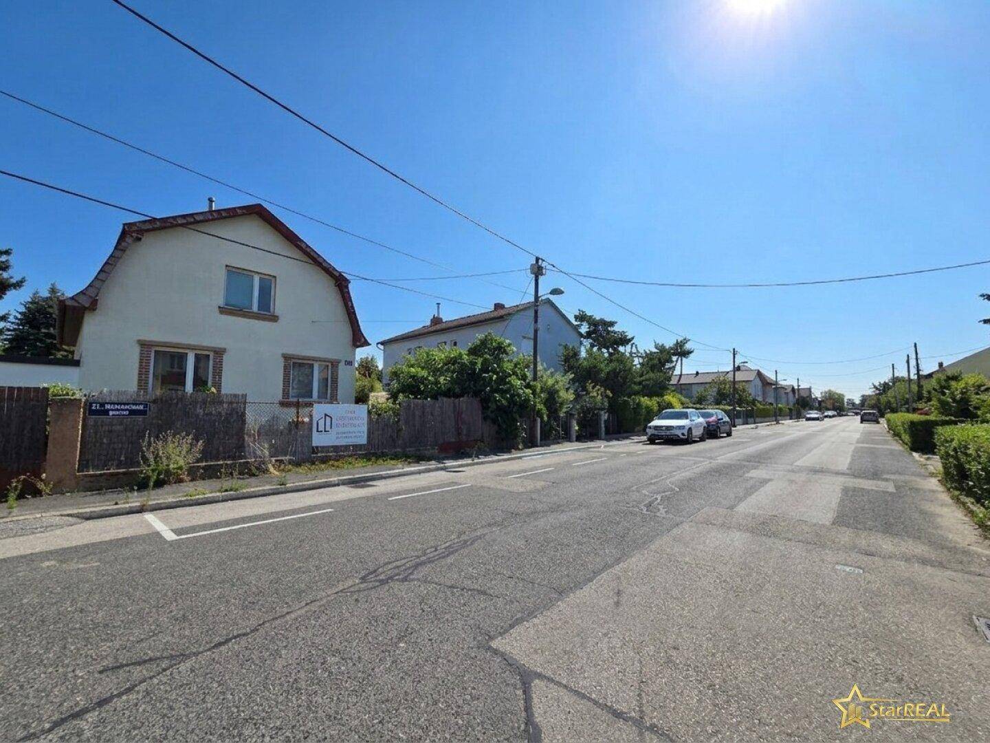 Weitläufige Ansicht einer sonnigen Wohnstraße mit mehreren Häusern, Bäumen und Hecken.