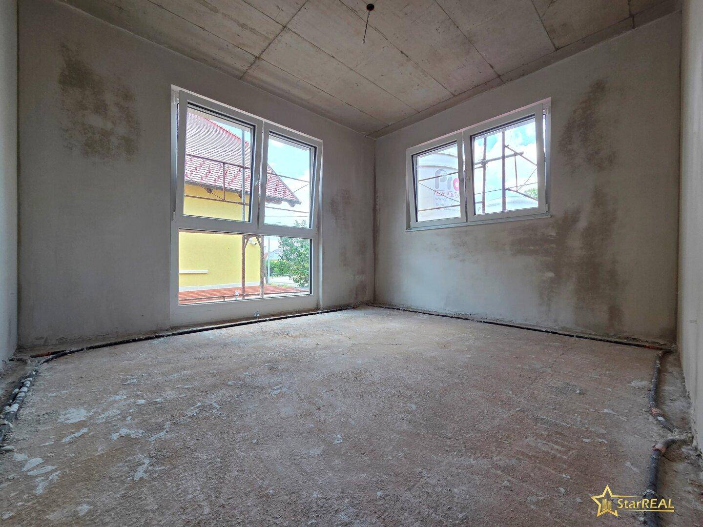 Unfertiger Raum mit großen Fenstern, die viel Tageslicht hereinlassen und Blick nach draußen bieten.