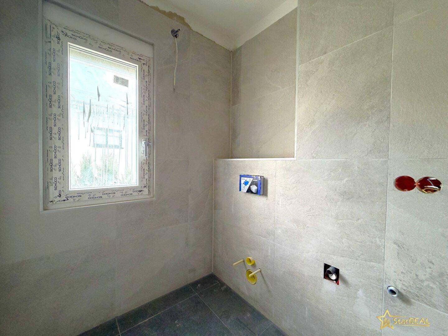 Unfertiges Badezimmer mit hellgrauen Fliesen, Fenster und Anschlüssen für Sanitäranlagen.