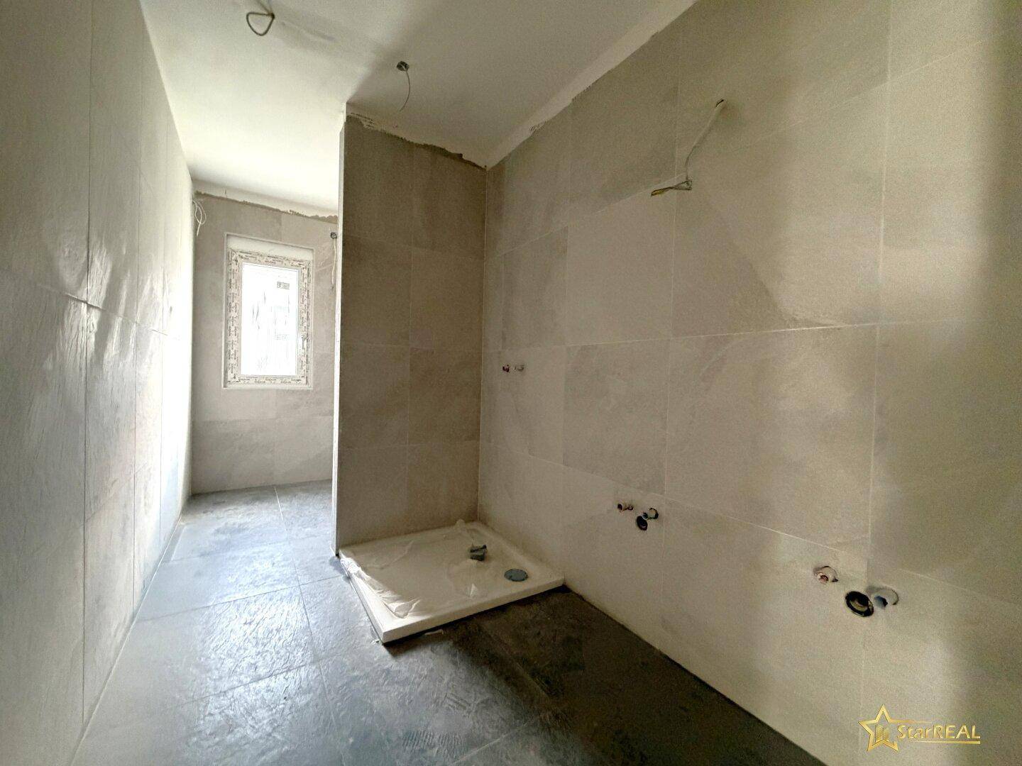 Badezimmer im Rohbau mit grauen Fliesen, Duschwanne und Fenster, bereit für die Installation.
