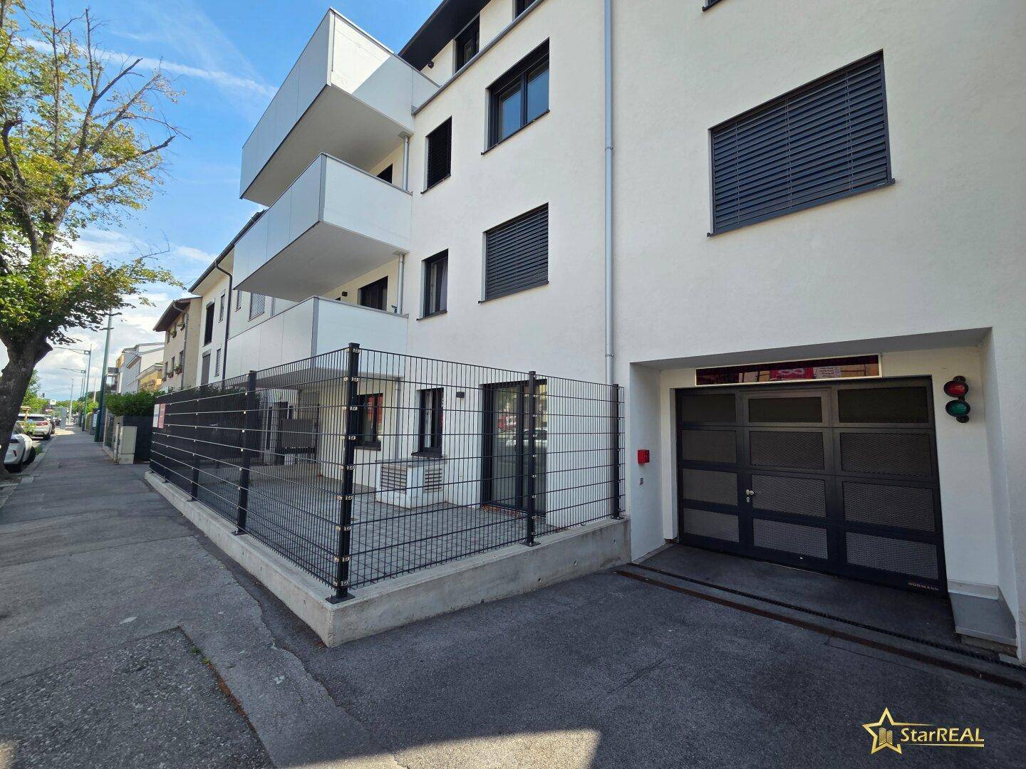 Seitenansicht des modernen Gebäudes mit Tiefgaragenzufahrt und umzäuntem Bereich im Erdgeschoss.