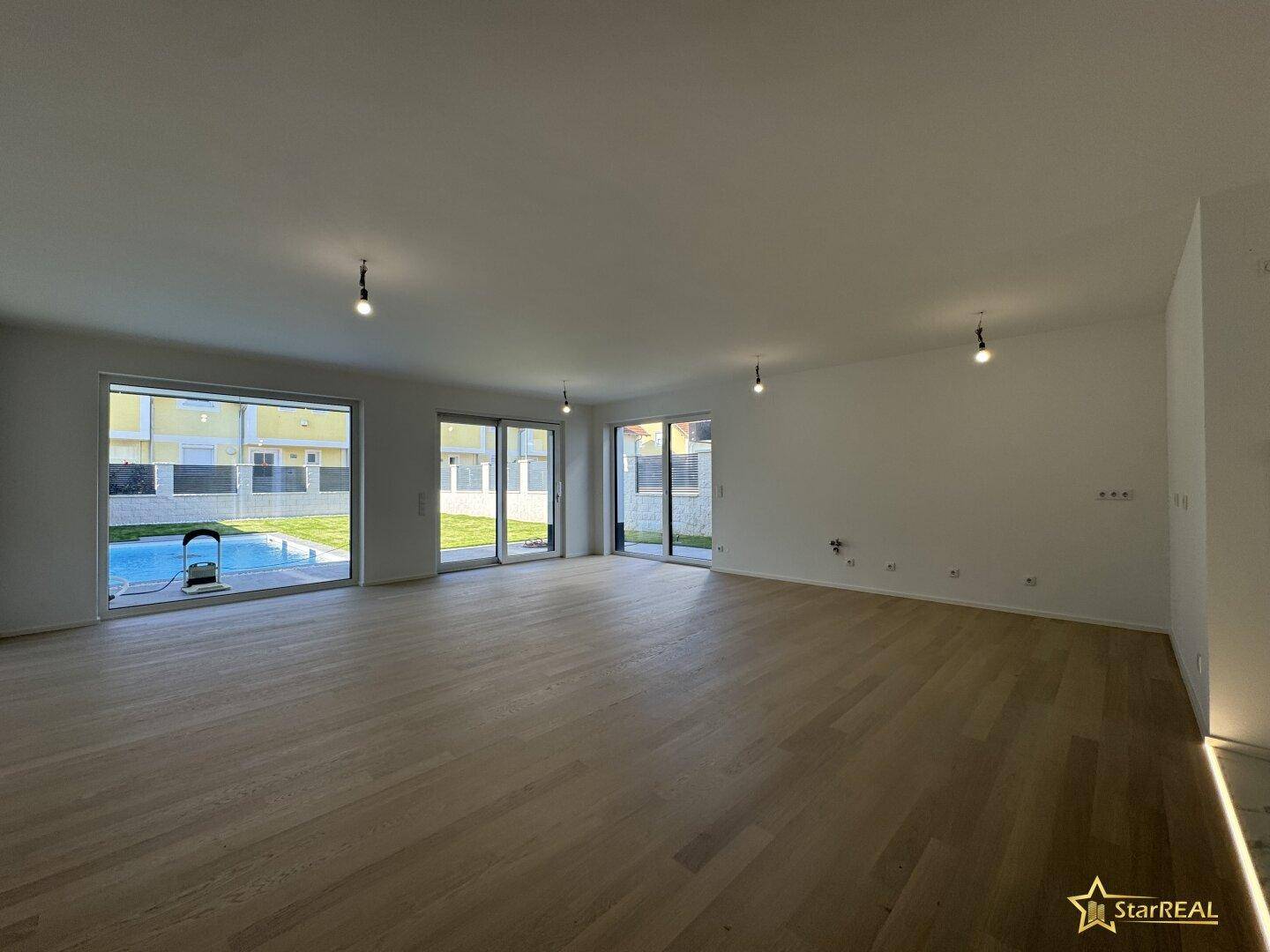 Großzügiges, leeres Wohnzimmer mit hellem Parkettboden und Zugang zum Garten mit Pool.