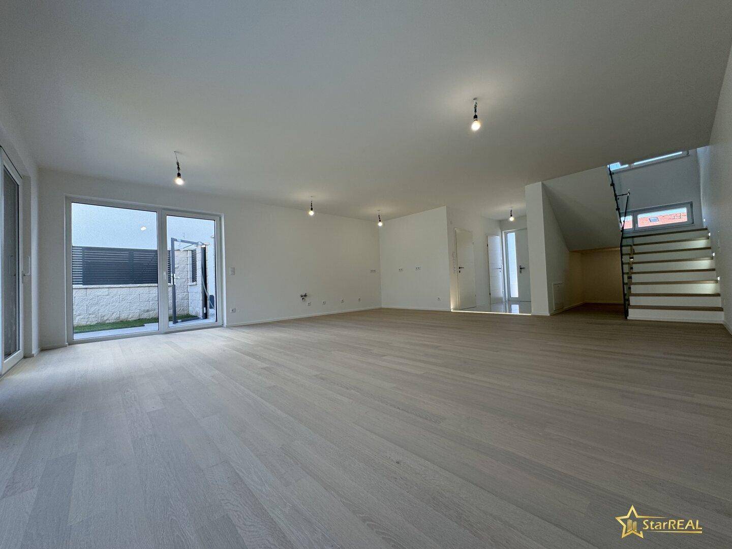 Weitläufiger Wohnbereich mit Parkettboden, großen Fensterfronten und Blick auf den Treppenaufgang.