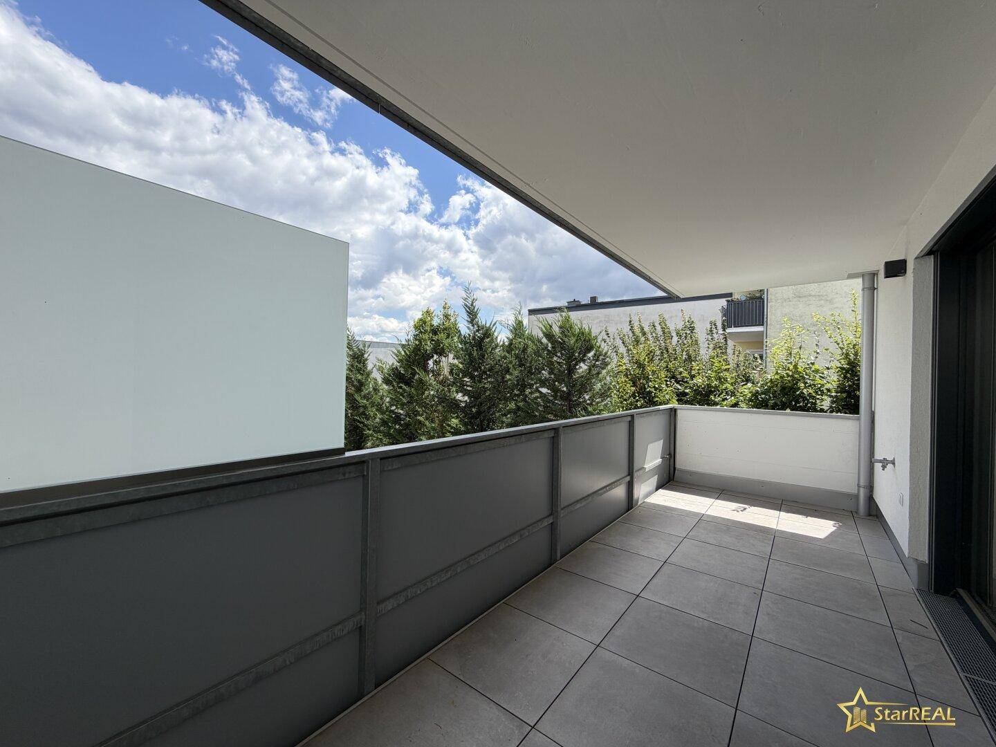 Geräumiger Balkon mit grauen Fliesen, moderner Brüstung und einem angenehmen Blick ins Grüne.