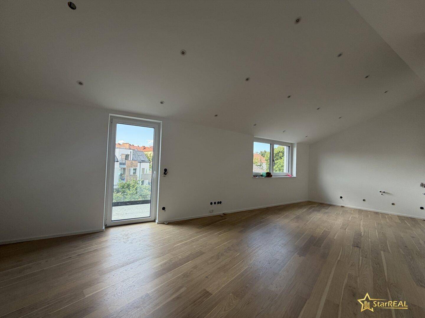 Heller, leerer Raum mit Holzboden, weißen Wänden und Zugang zu einem Außenbereich.