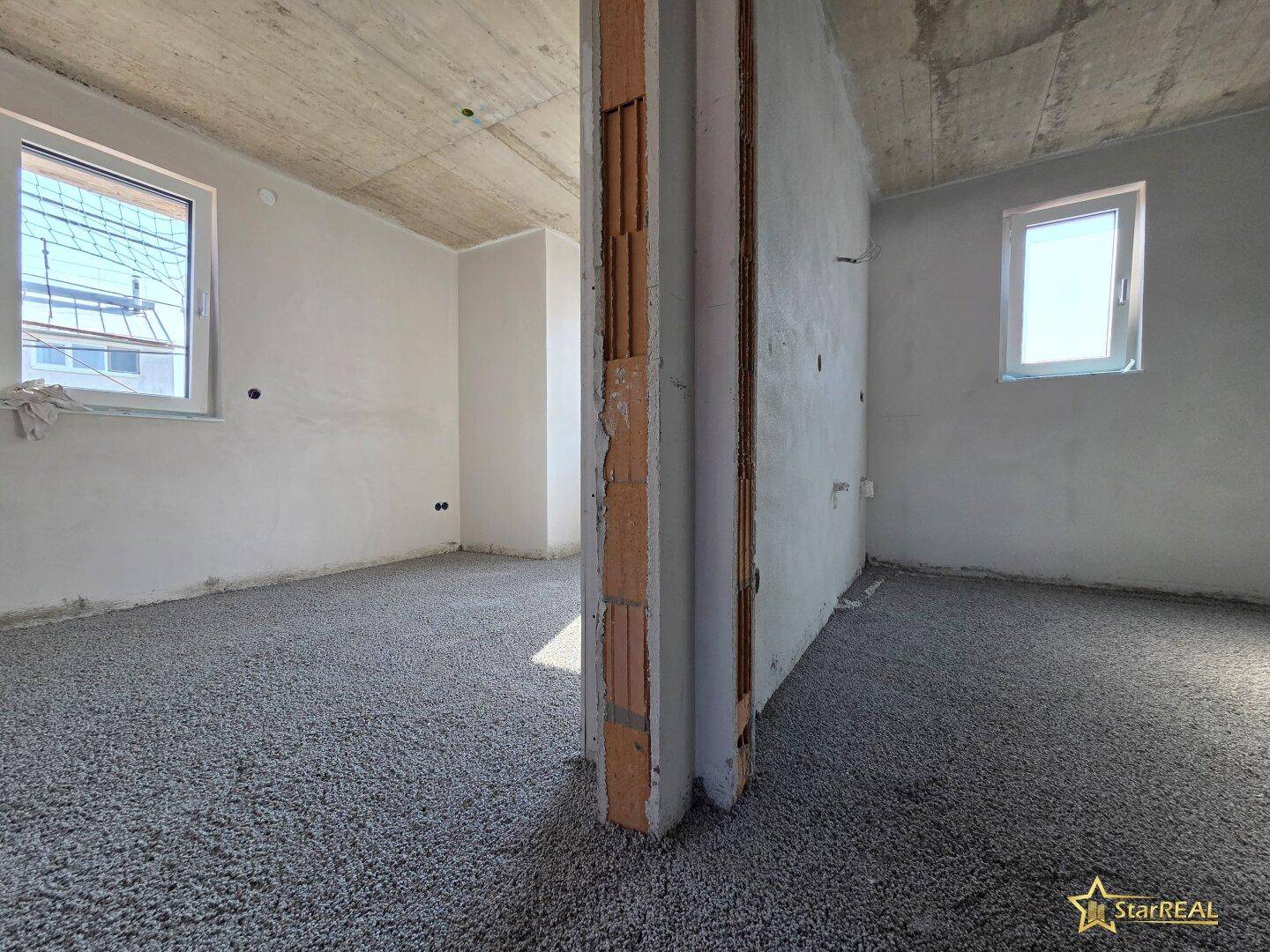 Rohbau-Raum mit teilweise errichteter Wand und zwei Fenstern, viel Tageslicht.