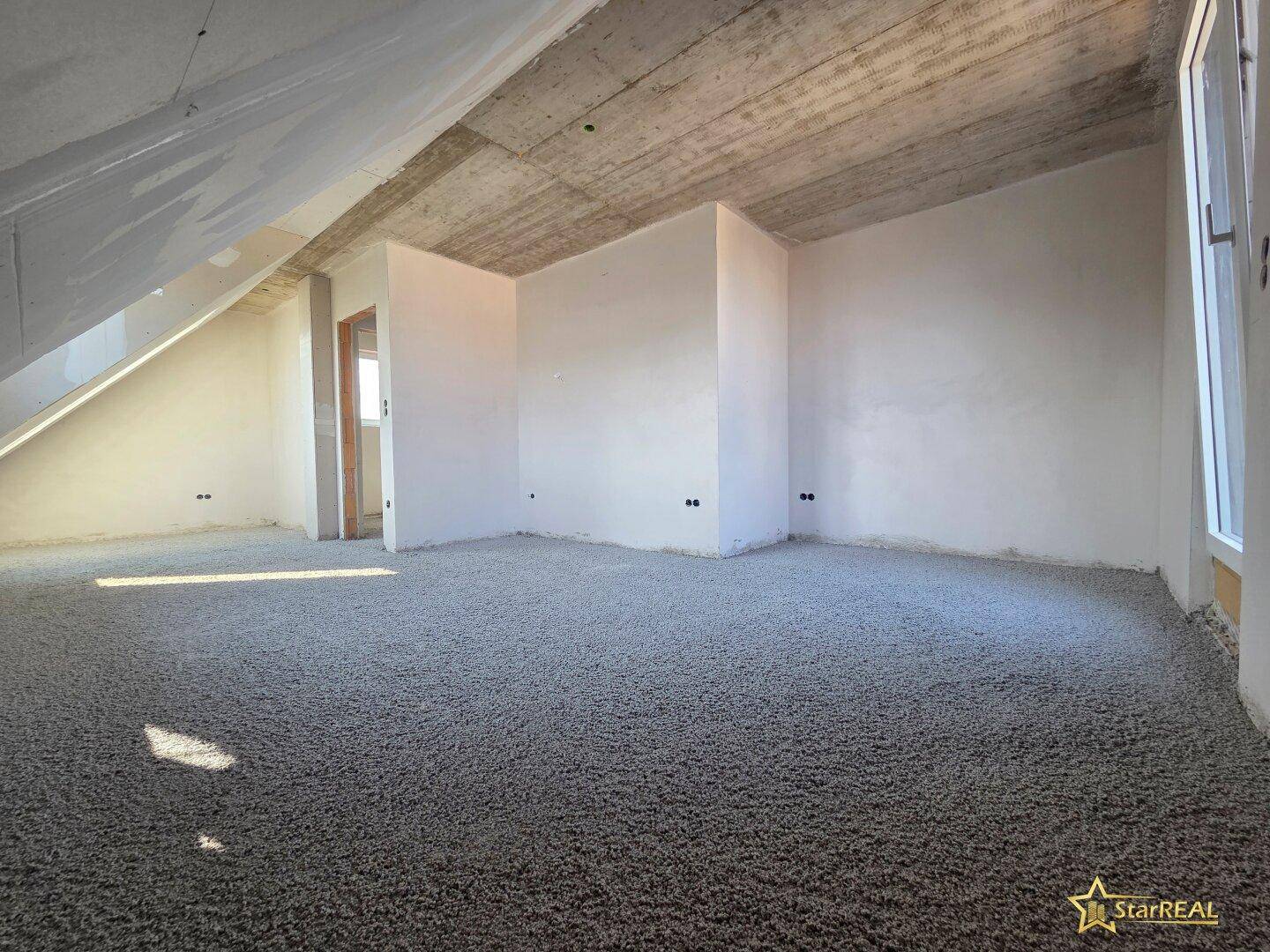 Dachgeschossraum im Rohbau mit schräger Decke und Fenster, viel natürliches Licht.