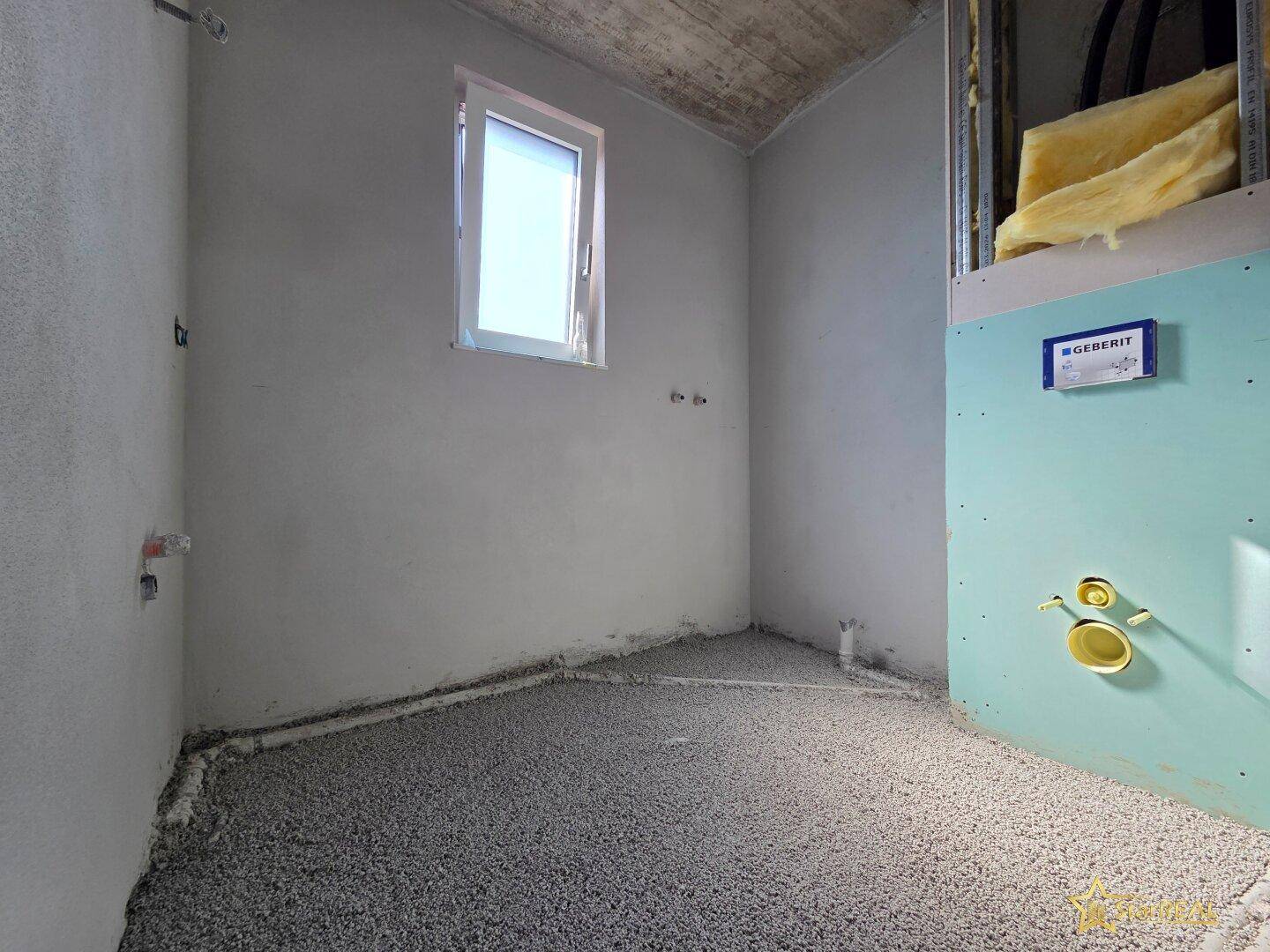 Rohbauraum mit sichtbaren Sanitärinstallationen für ein WC, einem Fenster und Dämmmaterial.