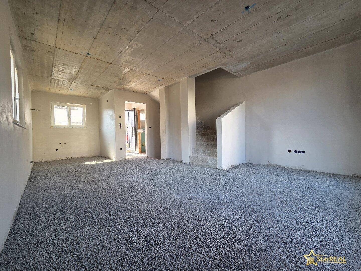 Großer Raum im Rohbau mit Beton-Decke, unfertigem Boden und sichtbarer Treppe.