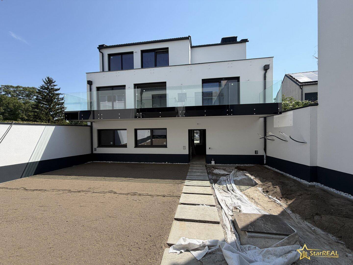 Neubau-Fassade mit großzügiger Terrasse und vorbereitetem Gartenbereich im Erdgeschoss an einem sonnigen Tag.