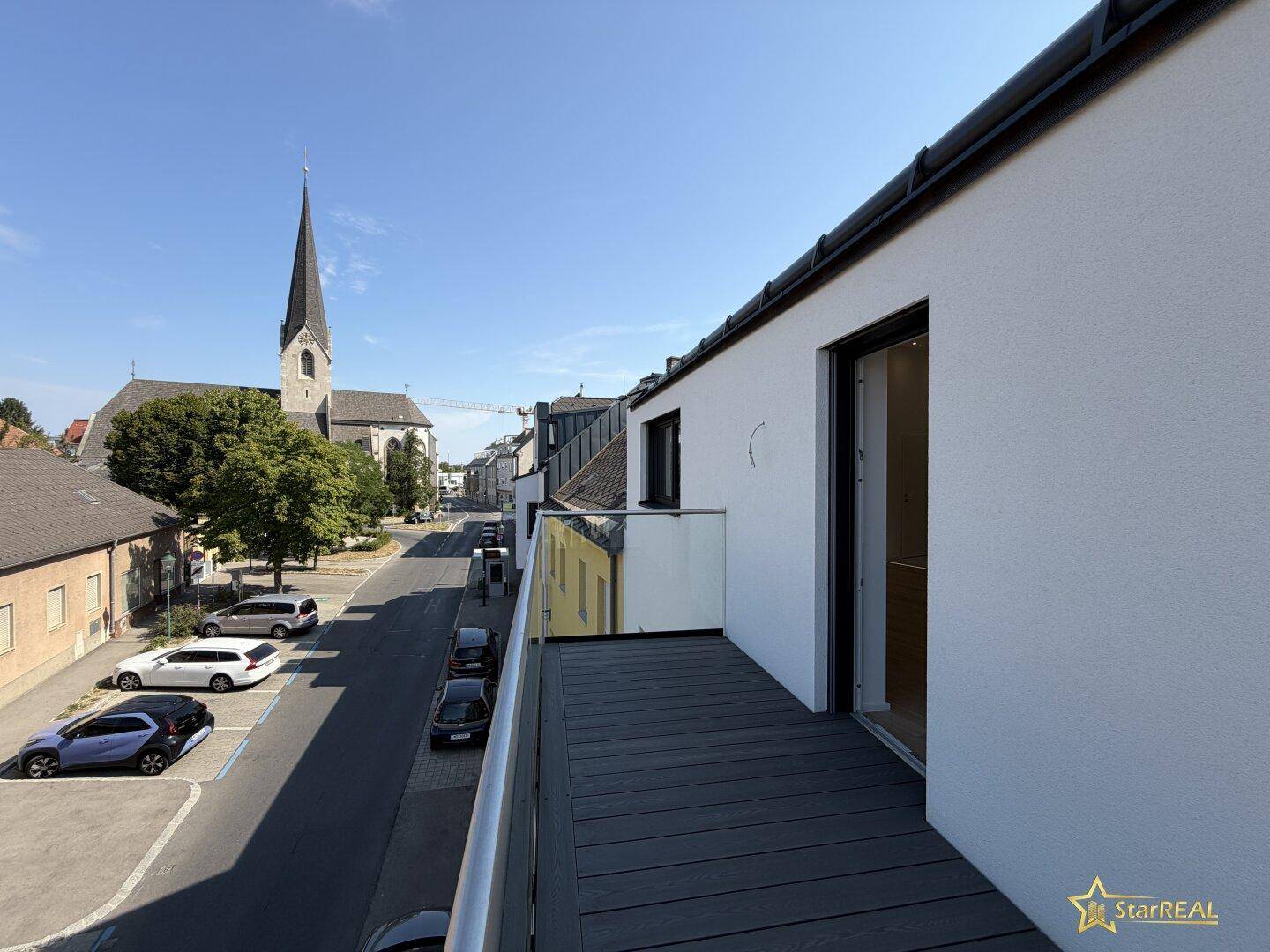 Langer Balkon mit Holzbelag und Glasgeländer, bietet einen Blick auf die Straße und eine Kirche.