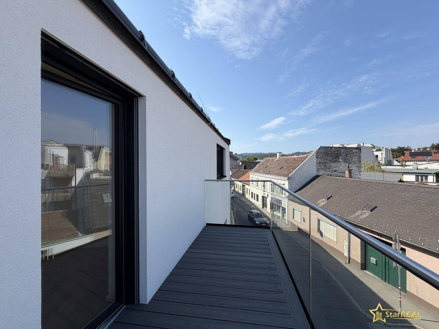 Schmaler Balkon mit modernem Holzbelag und Glasgeländer, mit Blick auf die Dächer der Stadt.