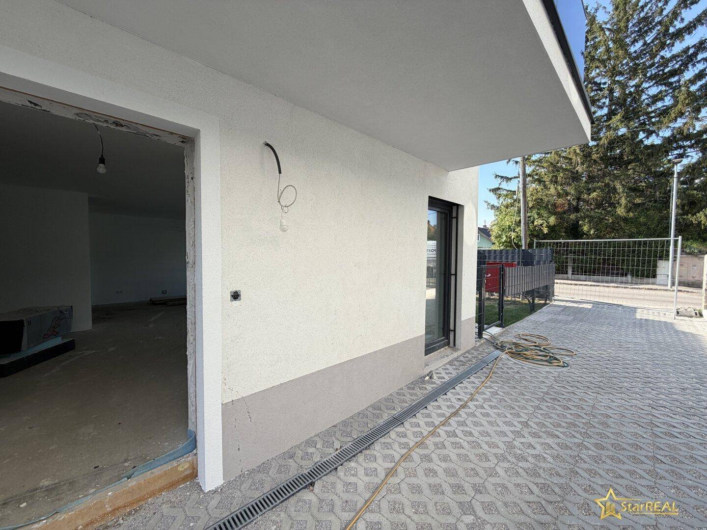 Seitenansicht des Neubaus mit gepflasterter Zufahrt und offener Tür zu einem unfertigen Innenraum.