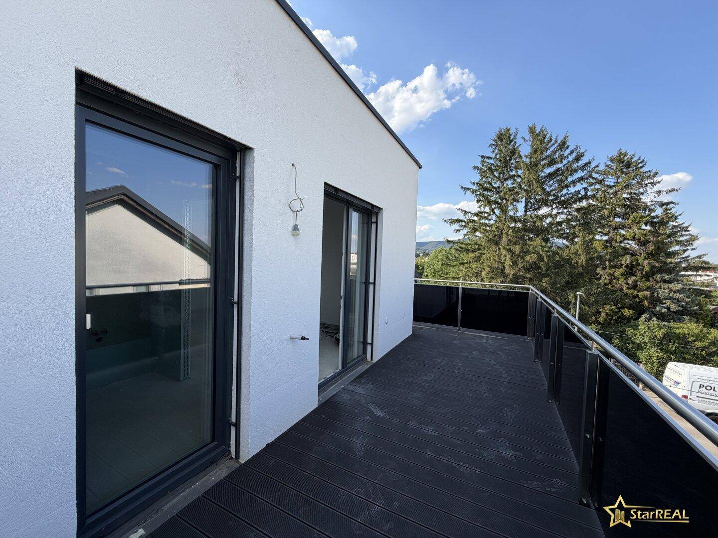 Geräumiger Balkon mit dunklem Holzboden und Glasgeländer, umgeben von Bäumen.