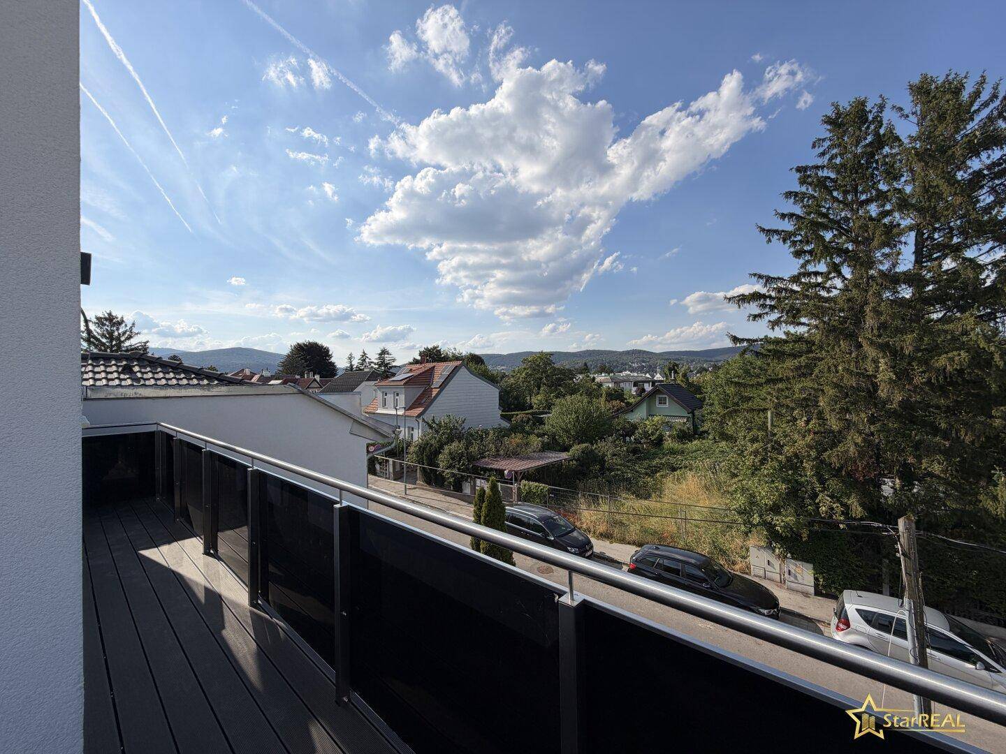 Balkon mit Blick auf die umliegende Wohngegend und grüne Bäume unter blauem Himmel.