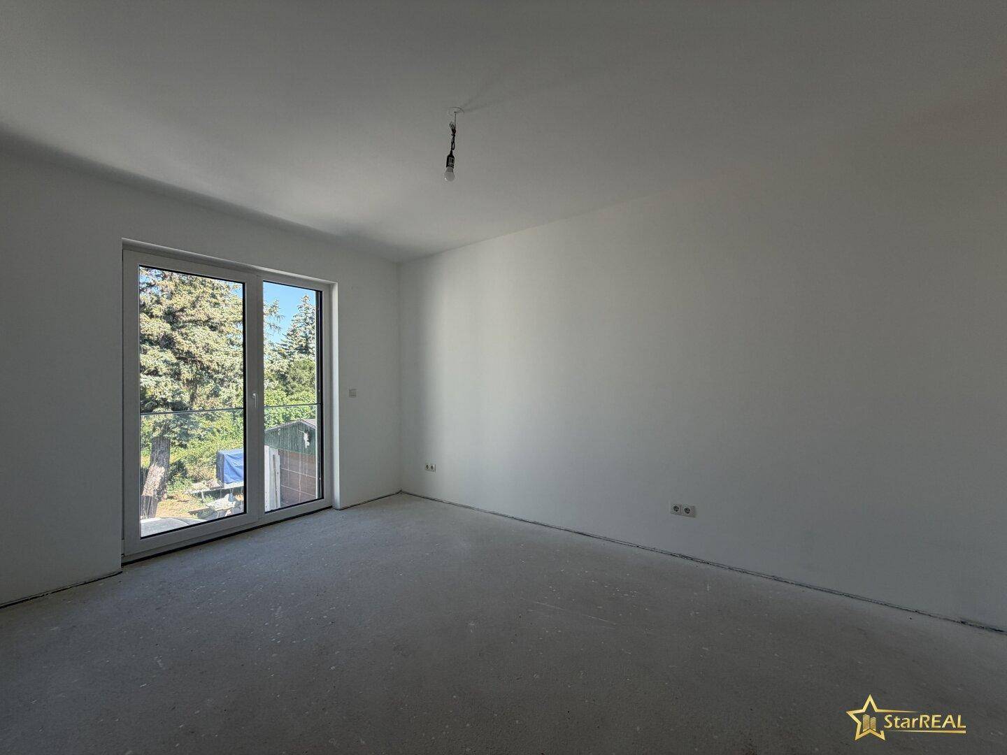 Unmöbliertes Zimmer mit großem Fenster, das viel Tageslicht und eine grüne Aussicht bietet.