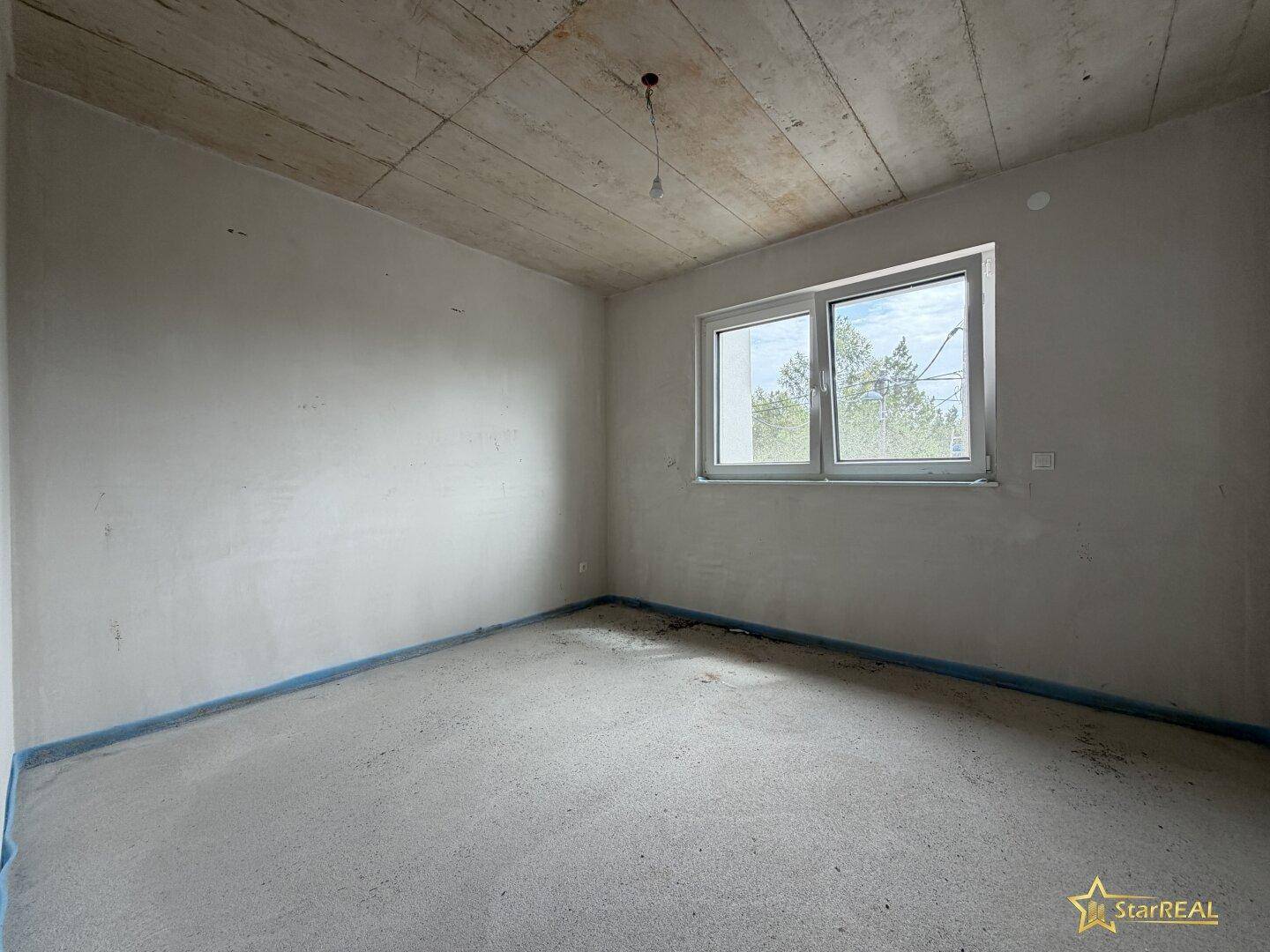 Leerer Raum im Rohbau mit Betonboden und einem großen Fenster, ideal für individuelle Gestaltung.