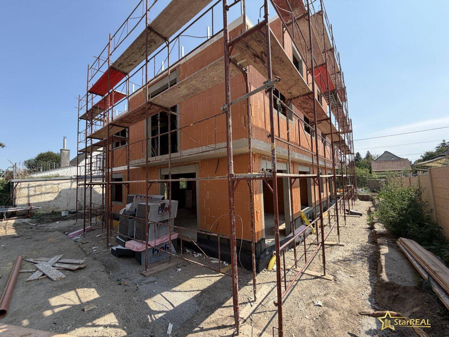 Neubau im Rohbau mit Gerüst und Ziegelmauerwerk in sonniger Umgebung.