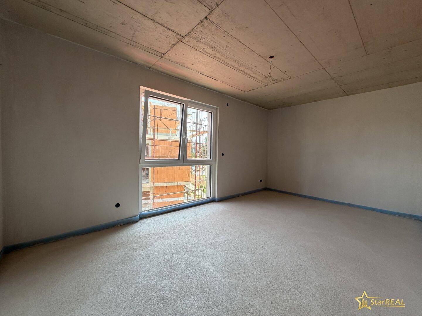 Schlafzimmer im Rohbau mit großem Fenster und Blick auf die umliegende Baustelle.