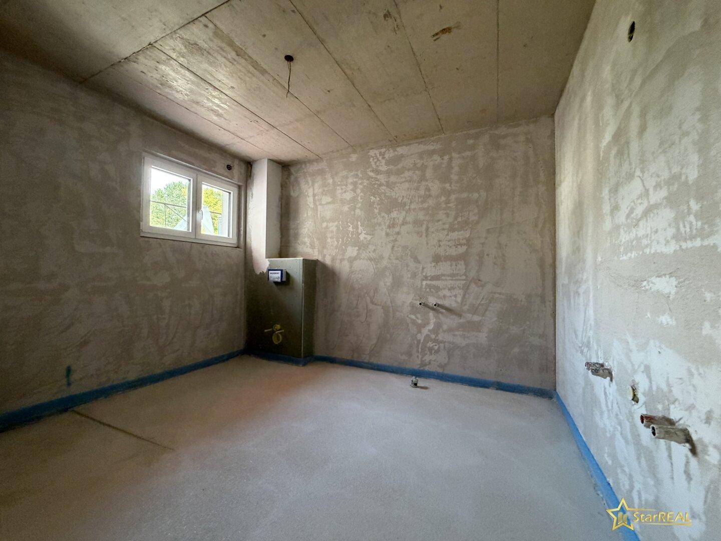 Rohbauzimmer mit Fenster, sichtbaren Installationen und Betonboden.