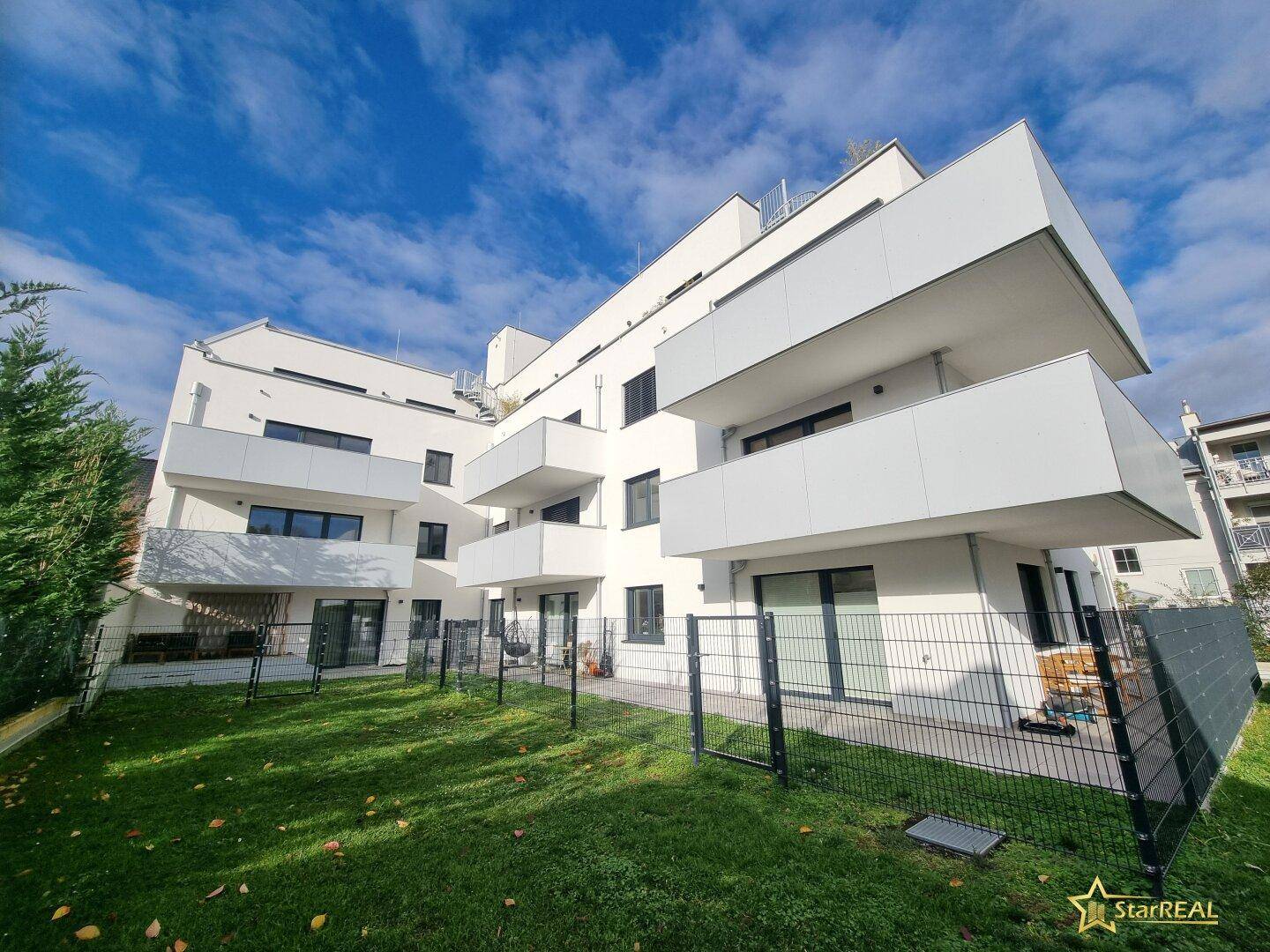 Modernes Wohngebäude mit weißer Fassade, mehreren Balkonen und gepflegter Grünfläche unter blauem Himmel.