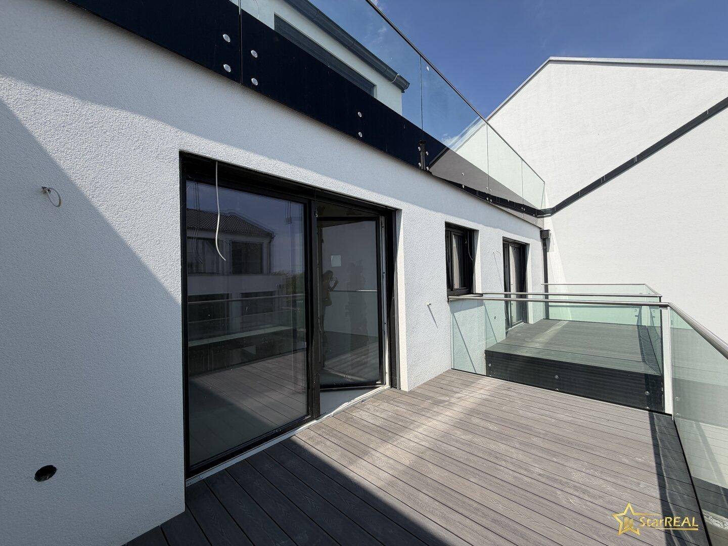 Geräumiger Balkon mit Holzdielen und Glasgeländer, zugänglich durch große Schiebetüren.