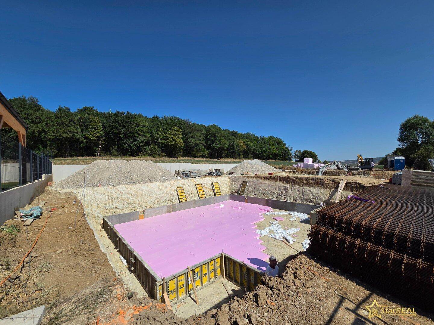 Baustelle mit ausgehobener Baugrube und vorbereitetem Fundament für ein neues Gebäude unter blauem Himmel.