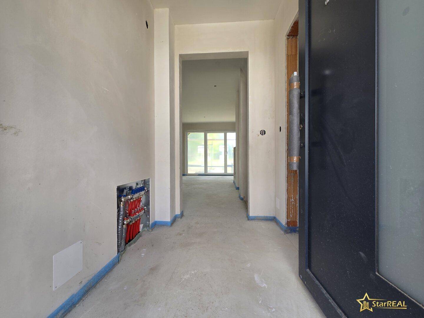 Eingangsbereich im Rohbau mit sichtbaren Installationen und Blick in einen hellen Raum.