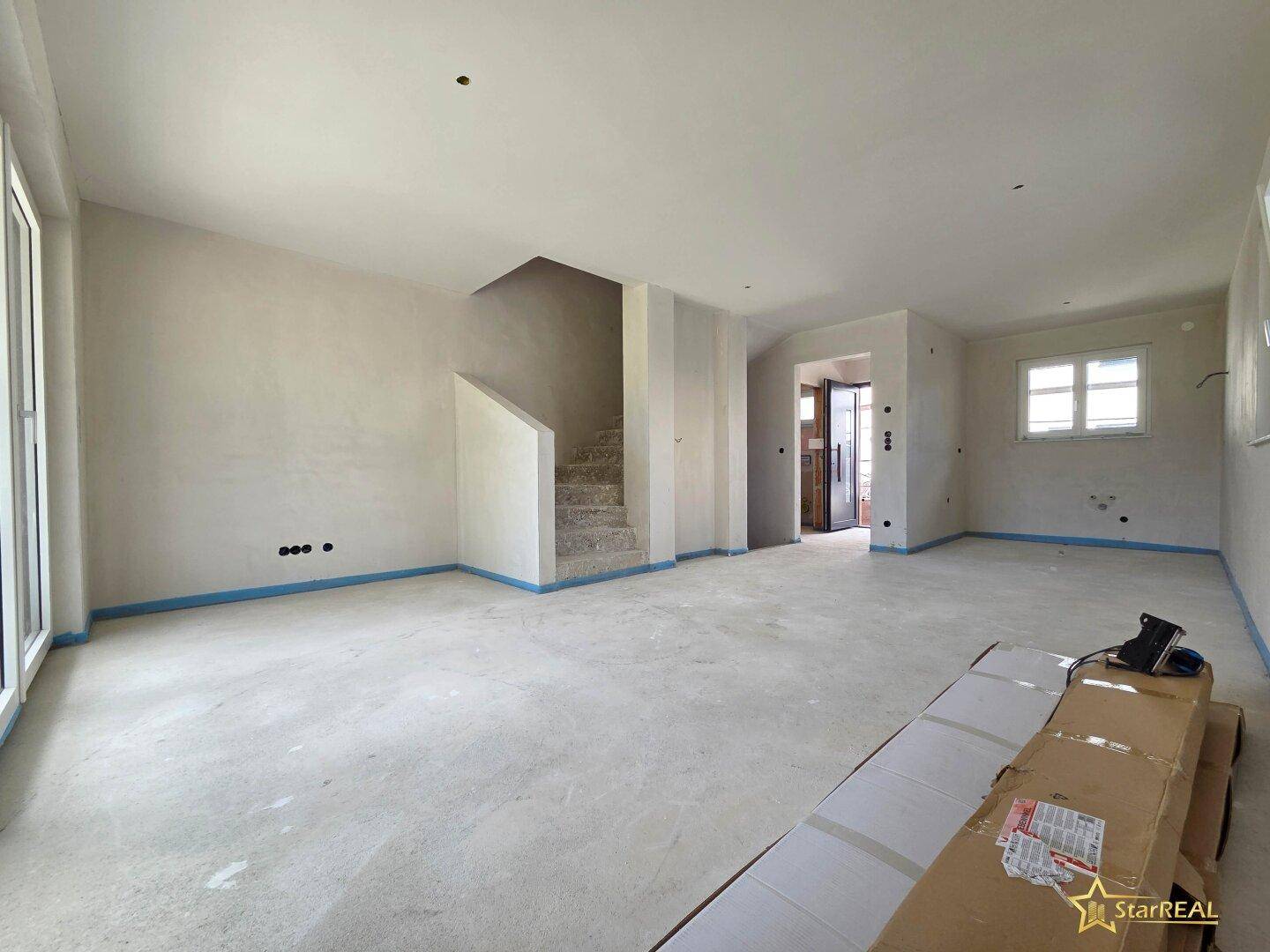 Großer Raum im Rohbau mit Treppenaufgang und vorbereiteten Wänden für den Innenausbau.