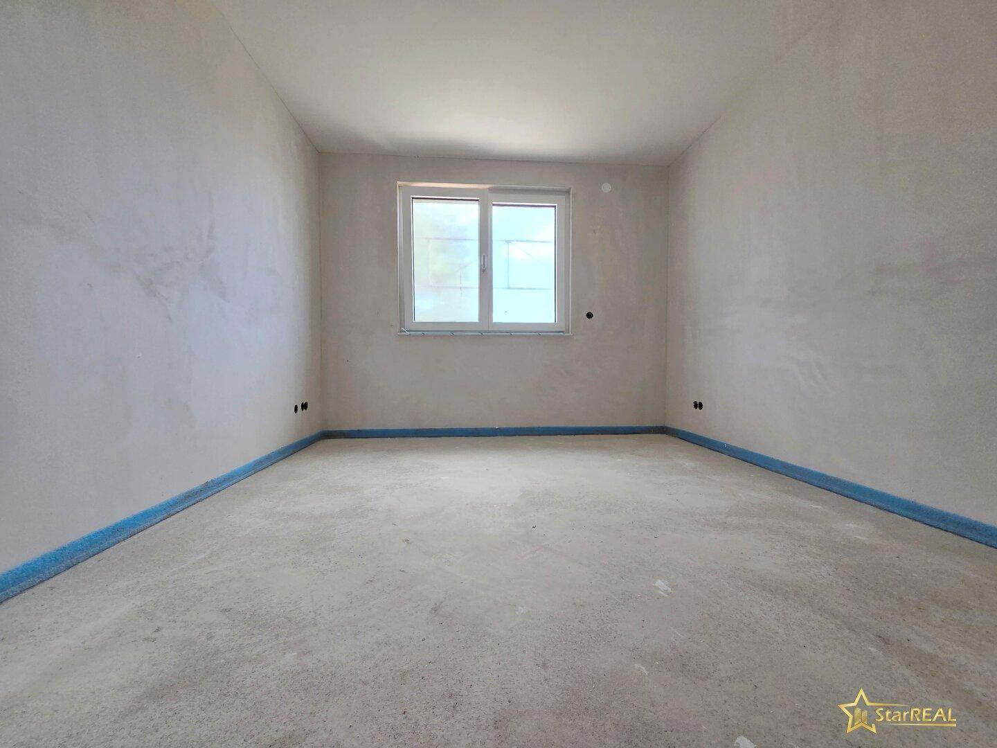 Zimmer im Rohbau mit einem Fenster, das Tageslicht hereinlässt, und unfertigem Boden.