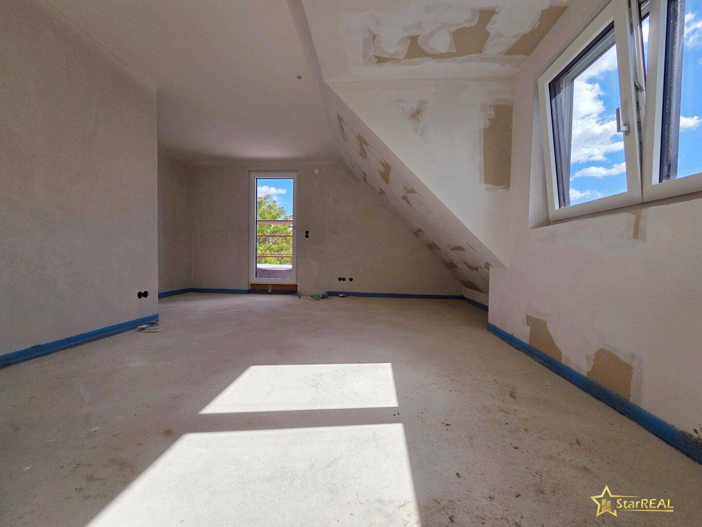 Dachgeschosszimmer im Rohbau mit schrägen Wänden, Fenster und viel natürlichem Licht.