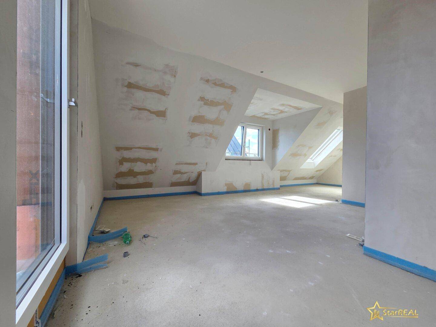 Dachgeschosszimmer im Rohbau mit schrägen Wänden, Gaubenfenster und Dachfenstern für Helligkeit.