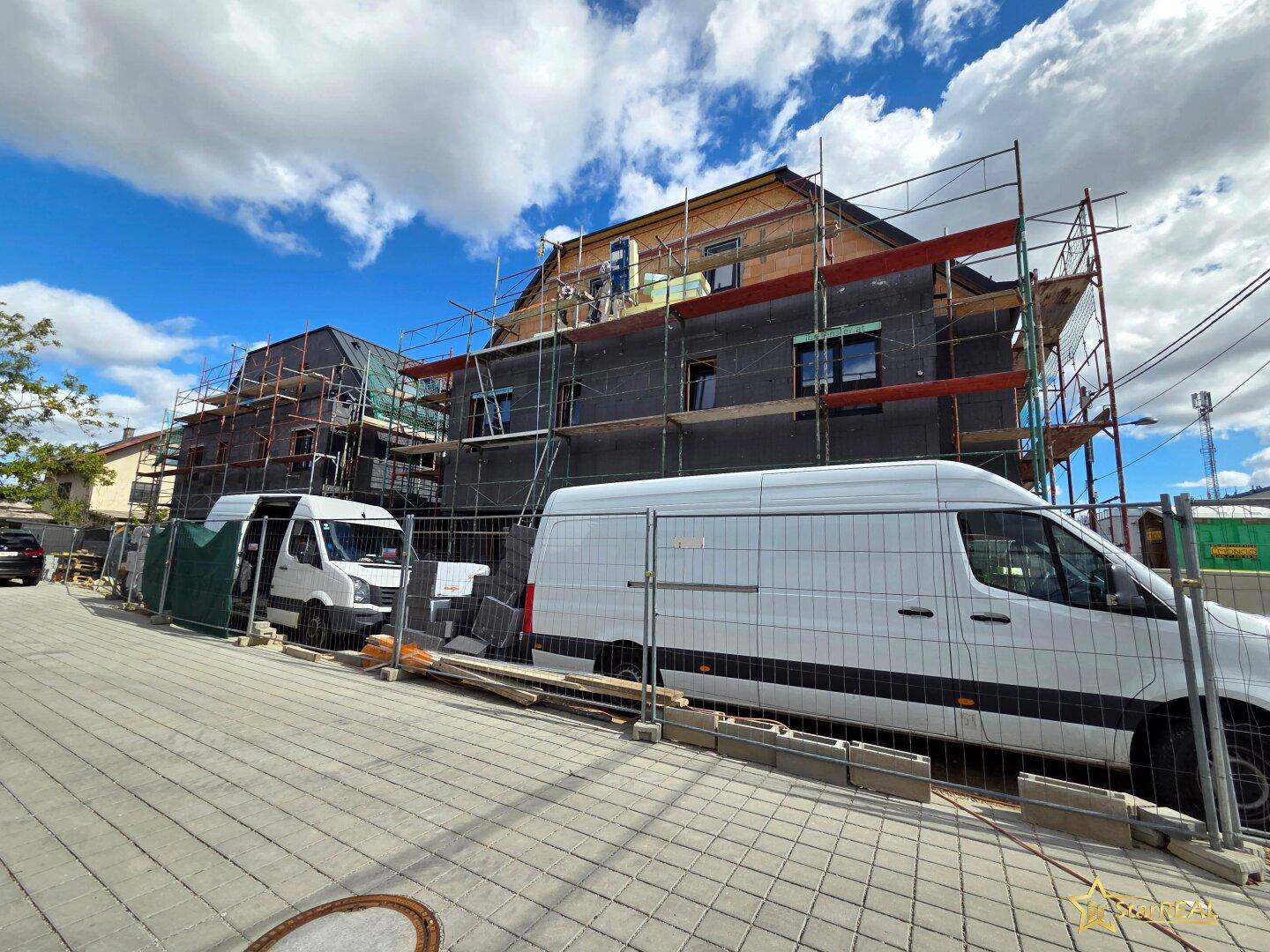 Straßenansicht eines Neubaus im Bauzustand mit Gerüst und parkenden Lieferwagen.