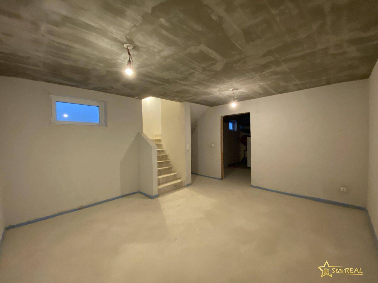 Heller Kellerraum im Rohbau mit Betonboden, kleinem Fenster und Zugang über eine Treppe.