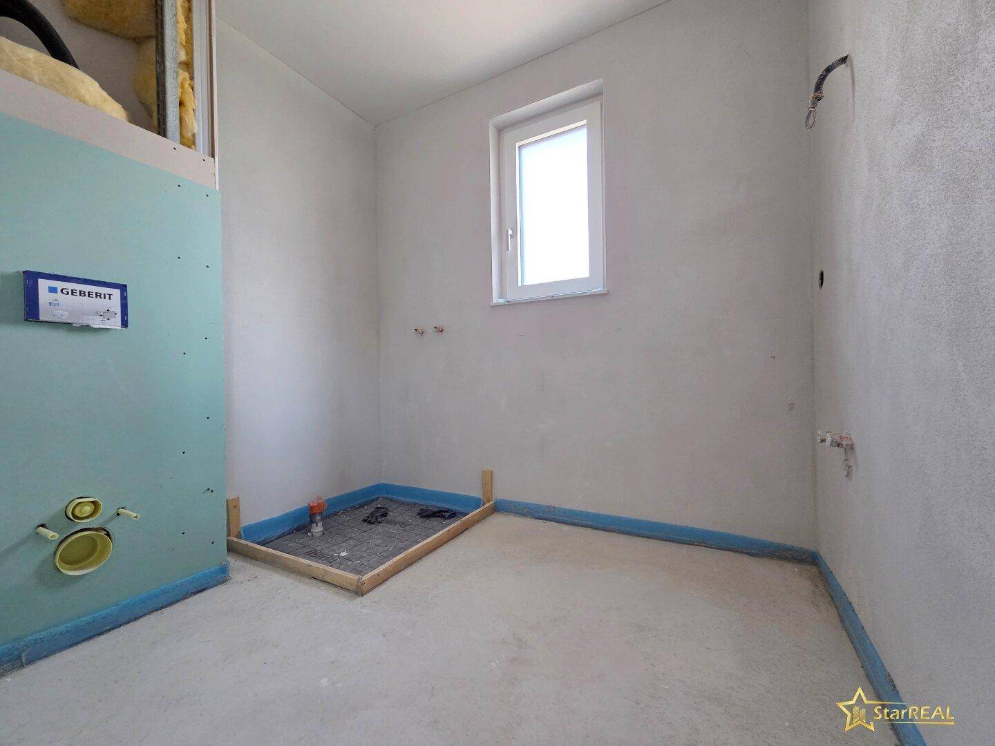 Badezimmer im Rohbau mit vorbereiteter Duschwanne und Fenster für natürliche Belüftung.