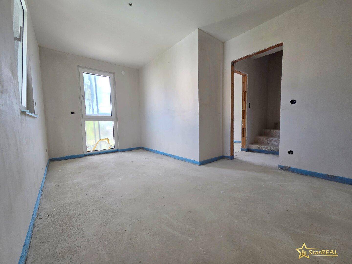 Unfertiges Zimmer mit Fenster und Tür nach draußen, im Rohbauzustand.