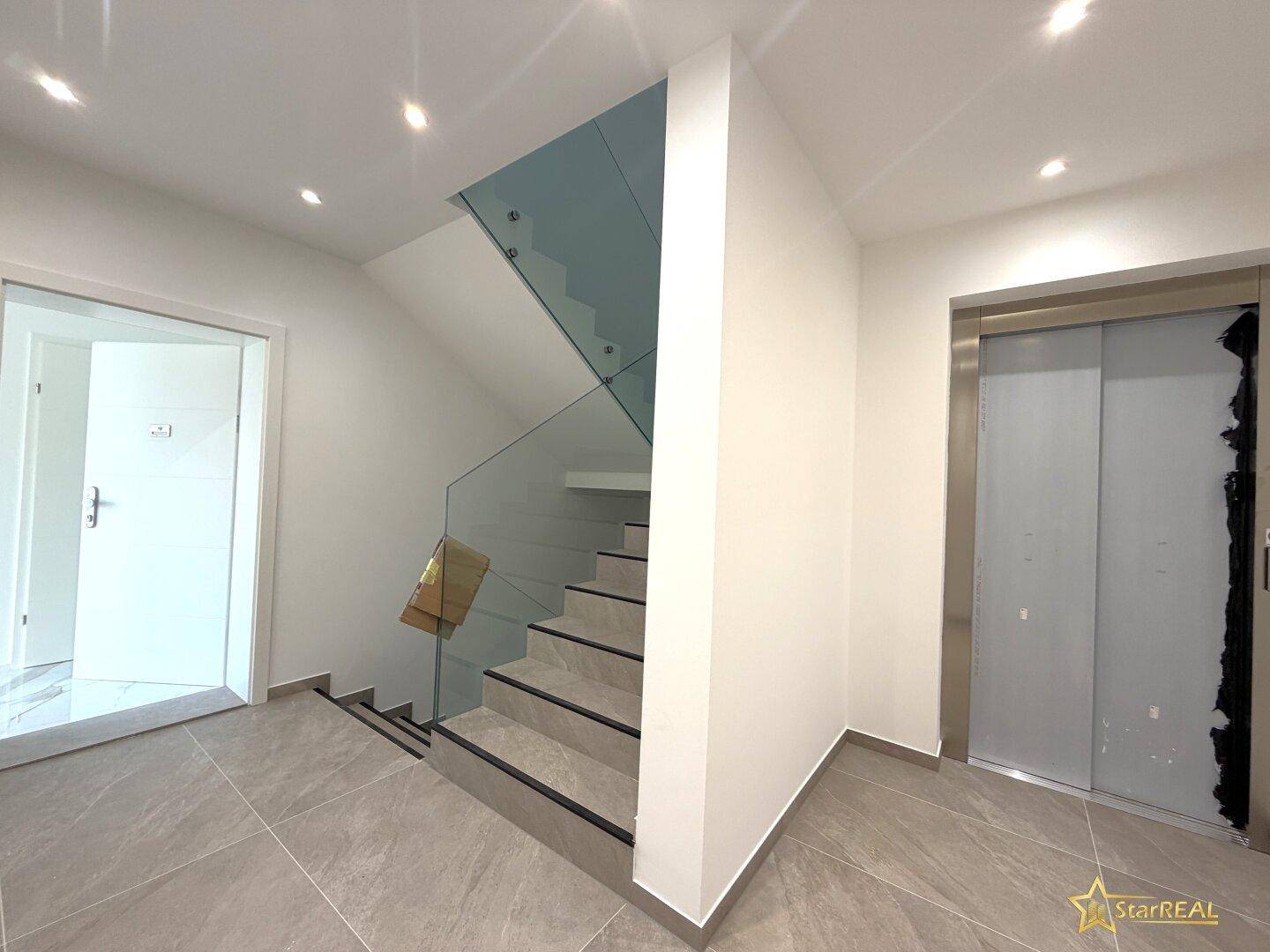 Moderner Eingangsbereich mit Treppe, Glasgeländer und Aufzug.