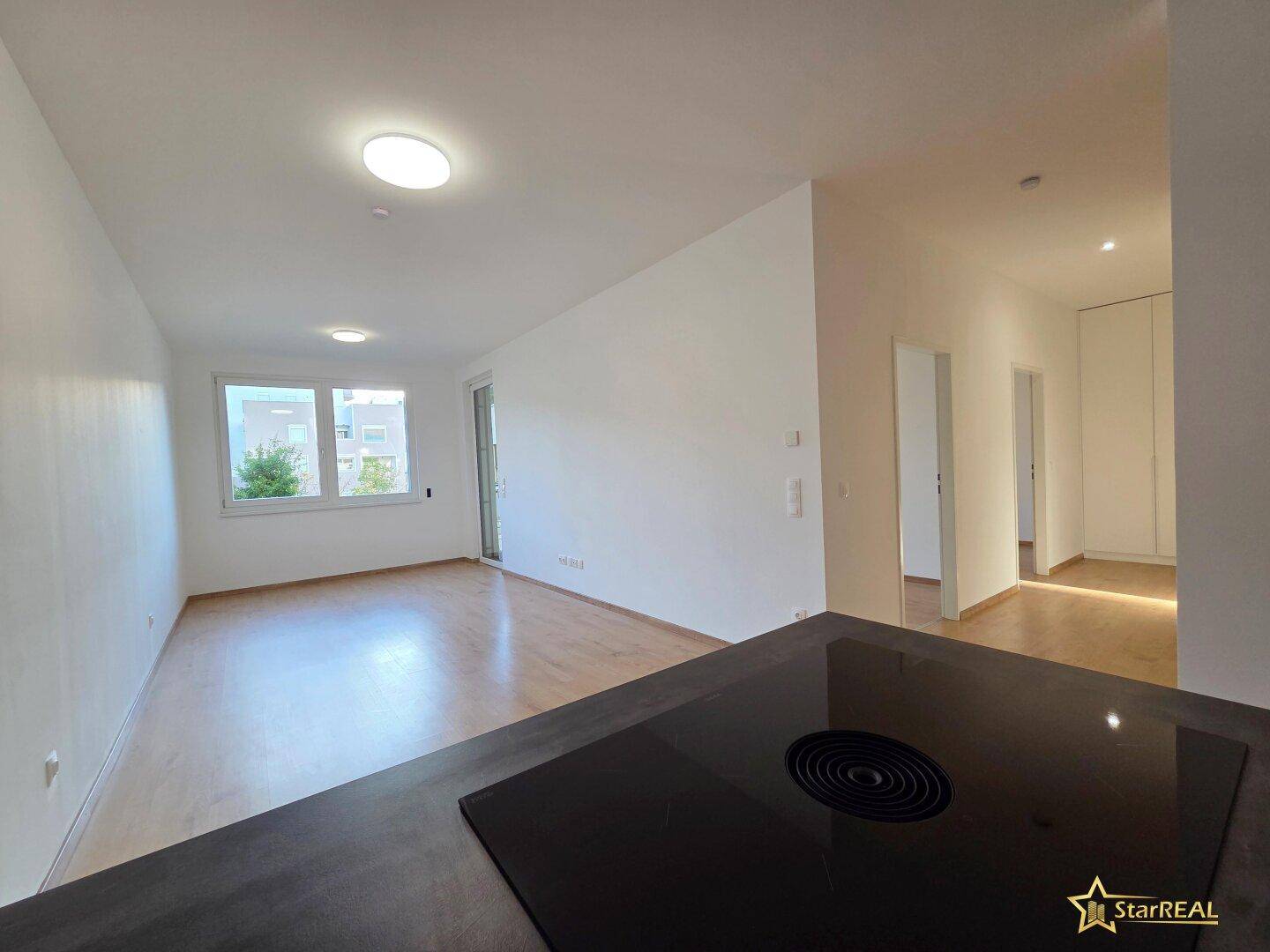 Heller, offener Wohnbereich mit Laminatboden und Blick auf eine moderne Küchenzeile mit Induktionskochfeld.