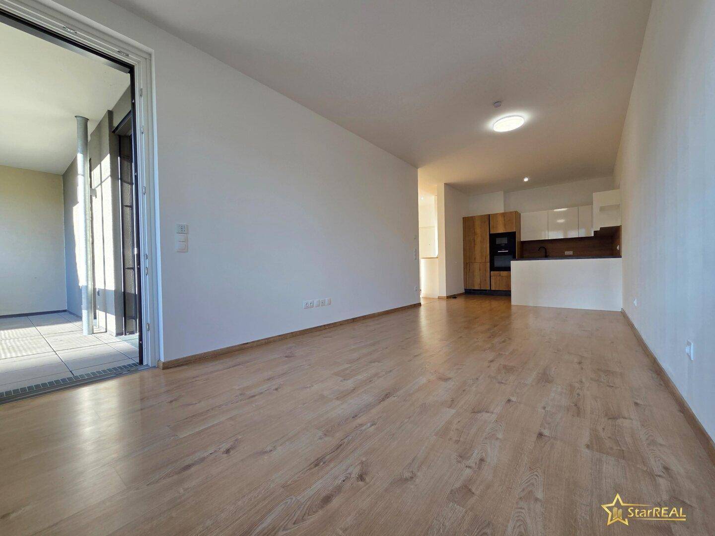 Heller Wohnbereich mit Laminatboden und Zugang zu einem Balkon, im Hintergrund die moderne Küche.